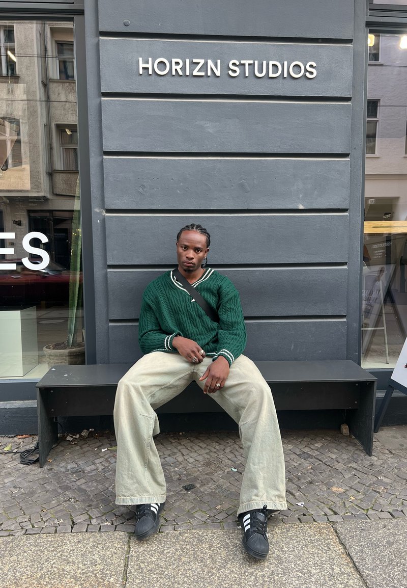 Πράσινο πουλόβερ με πλέξη καλωδίου και λευκές λεπτομέρειες, χαλαρές μπεζ παντελόνες και μαύρα αθλητικά παπούτσια. Καθισμένο σε ένα παγκάκι κοντά σε ένα κατάστημα.