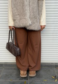 Gilet in pelliccia sintetica, top a maniche lunghe color crema, pantaloni marroni a gamba larga, stivaletti color cuoio alla caviglia e una borsa rettangolare marrone tenuta in mano.