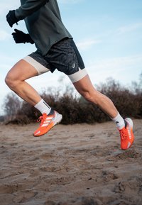 Figure athlétique dynamique en short de course et t-shirt, portant des chaussures d'entraînement orange vif et grises, sprintant sur un terrain sablonneux.