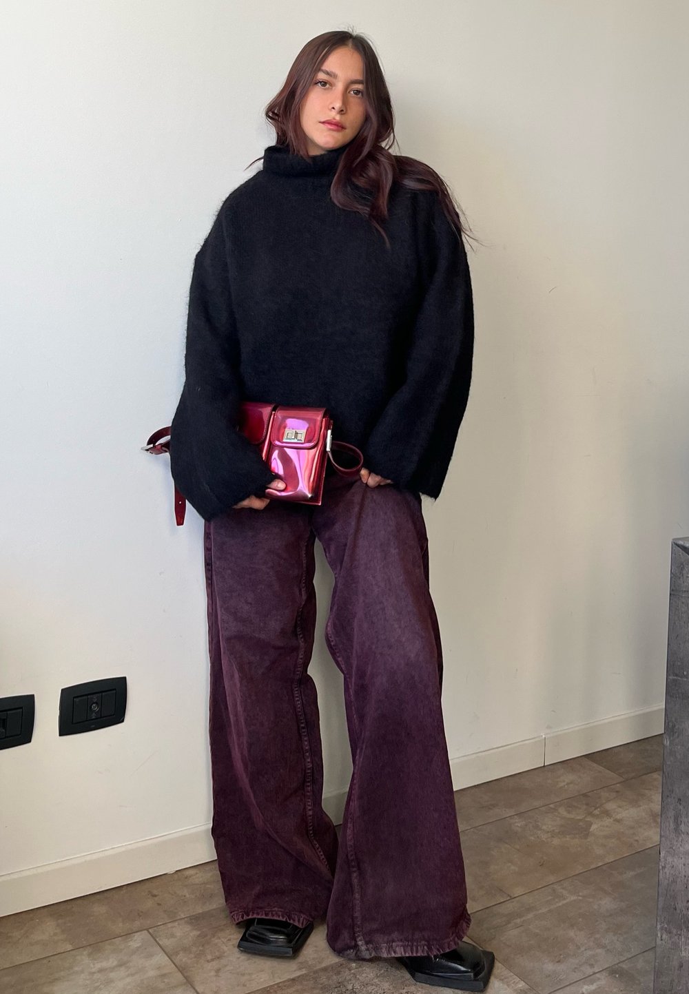 Överdimensionerad svart turtlenecktröja, vida burgundbyxor, blänkande rosa handväska och svarta fyrkantiga skor mot en ljus vägg.