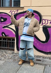 Μπεζ φλις μπουφάν, λευκό oversized πουκάμισο με μπλε ρίγες, φθαρμένα ανοιχτό μπλε τζιν και καφέ μποτάκια. Το φόντο περιλαμβάνει μωβ γκράφιτι.