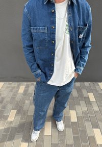 Blaues Denimhemd mit Vordertaschen über einem weißen T-Shirt mit Grafik, kombiniert mit locker sitzenden blauen Jeans und weißen Sneakers auf einem gemusterten Pflaster.