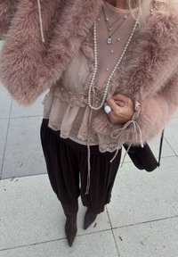 Person iført en lyserød fake pelsjakke, gennemsigtig blonde lyserød bluse, lagdelte perle- og guldkæder, sort nederdel og spidse mørke støvler på flisebelagt gulv.