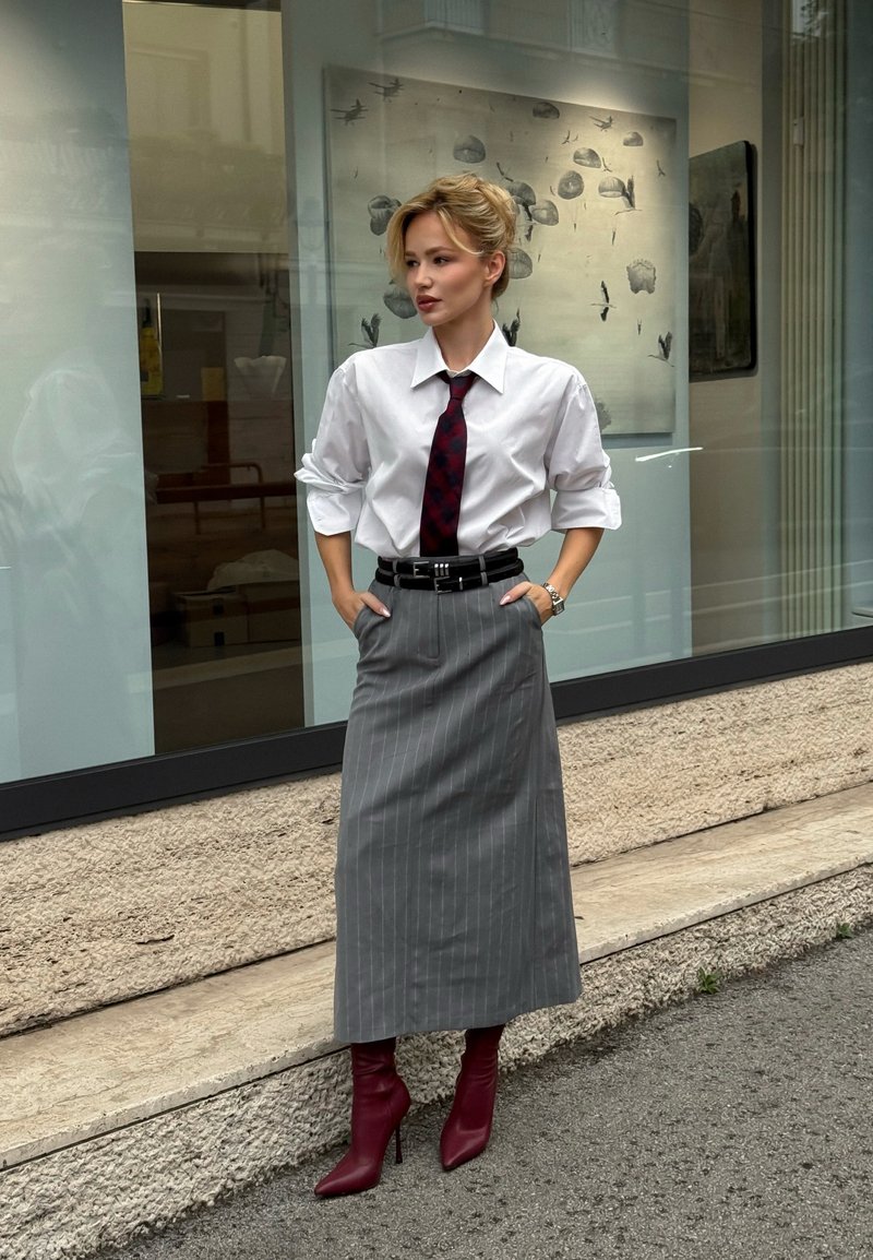 Žena stojí vonku oblečená v bielej košeli, červenej kravate, sivej sukni s tenkými pruhmi, čiernom opasku a červených členkových topánkach s podpätkom, ruky v kapsách.