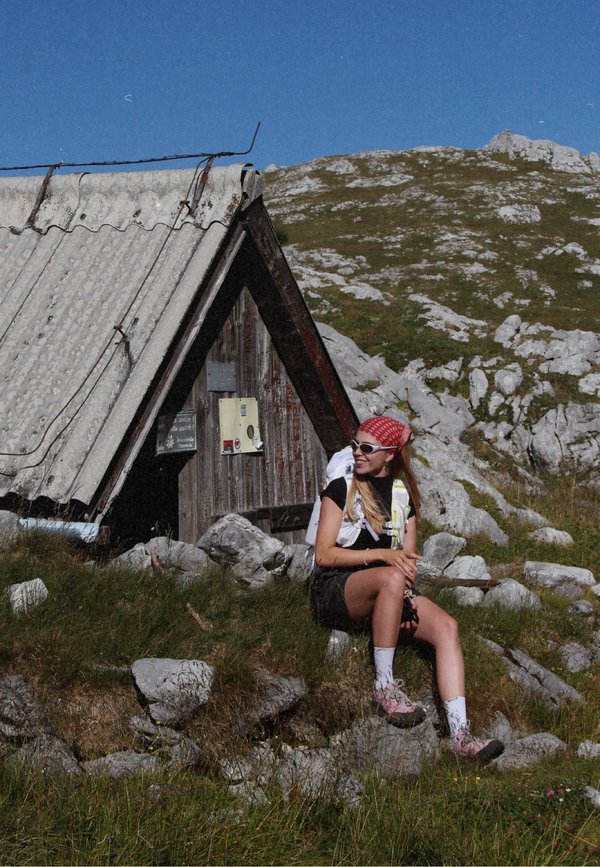 Jeune femme en tenue de randonnée assise sur des rochers près d'une petite cabane en bois de montagne sous un ciel bleu clair.