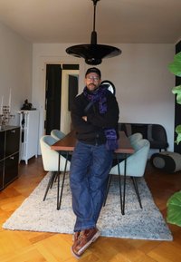 Mann med briller, caps og lilla skjerf som lener seg mot et spisebord i et moderne rom med stoler, teppe og hengelampe over.