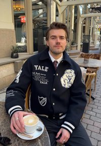 Casaco varsity preto com texto branco e um design de bulldog, usado sobre uma camisa branca e gravata, com uma chávena de café numa mesa.