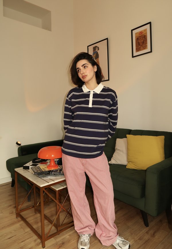 Polo a maniche lunghe a righe navy con colletto bianco; pantaloni a righe rosse e bianche; sneakers bianche casual; divano verde e lampada arancione sullo sfondo.