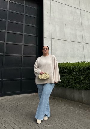 Suéter oversized beige claro con puños acanalados, combinado con jeans acampanados azul claro y un pequeño bolso amarillo. Fondo de una pared de concreto.