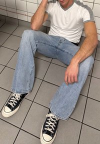 Άτομο που κάθεται σε πλακάκια δαπέδου, ακουμπώντας σε τοίχο με λευκά πλακάκια, φοράει λευκό και γκρι μπλουζάκι, ανοιχτό μπλε τζιν και μαύρα αθλητικά παπούτσια με λευκά κορδόνια.