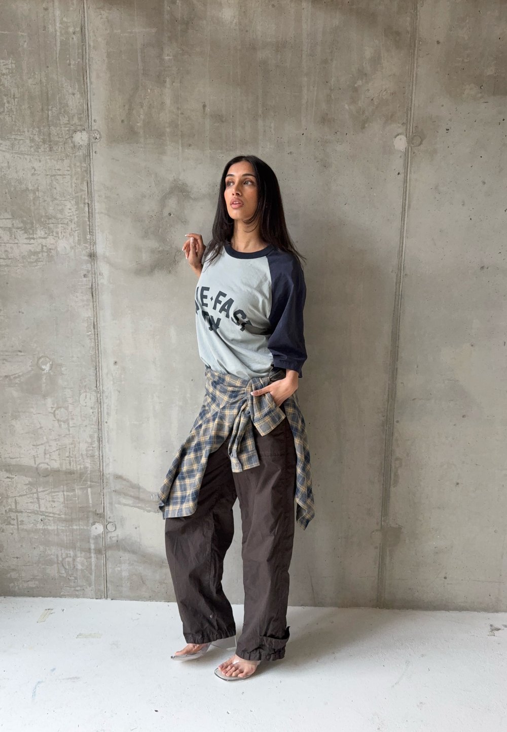 Femme debout contre un mur en béton, portant un t-shirt raglan graphique, une chemise à carreaux nouée à la taille, un pantalon ample foncé et des talons ouverts transparents.