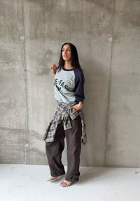 Femme debout contre un mur en béton, portant un t-shirt raglan graphique, une chemise à carreaux nouée à la taille, un pantalon ample foncé et des talons ouverts transparents.