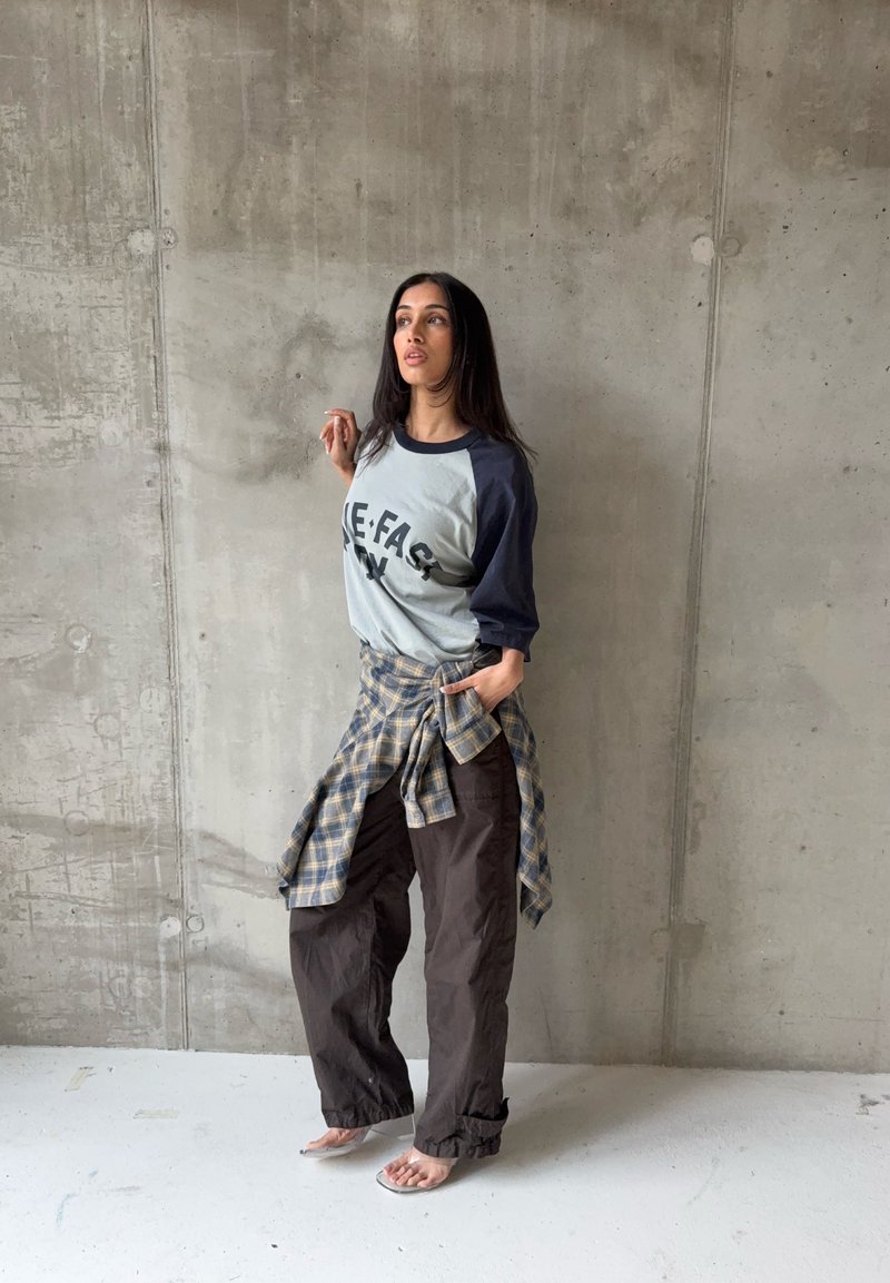 Femme debout contre un mur en béton, portant un t-shirt raglan graphique, une chemise à carreaux nouée à la taille, un pantalon ample foncé et des talons ouverts transparents.