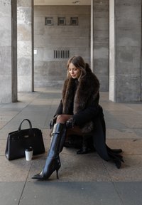 Čierne vysoké čižmy na ihličkovom podpätku po kolená, kabát s kožušinou a čierna kožená kabelka. Žena sediaca na chodníku, držiaca šálku kávy.