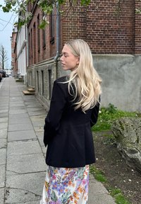 Blazer negro con corte entallado y solapas con muesca, combinado con una falda maxi de satén con un patrón floral que presenta colores vibrantes y acentos verdes.