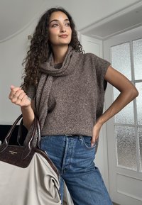 Brązowy sweter z krótkim rękawem i drapowanym dekoltem, w połączeniu z jasnoniebieskimi, wysokim stanem dżinsami. Nosi beżową torebkę z ciemnobrązowymi akcentami.