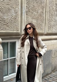 Μπεζ παλτό trench, σκούρα καφέ πουλόβερ, μαύρη φούστα, γυαλιά ηλίου, μαύρη τσάντα χειρός. Υπογείος τοίχος και παράθυρο στο φόντο.