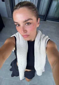 Fiatal nő, simára fésült hajjal, selfie-t készít, fekete crop topot, fekete szoknyát és a vállán drapírozott fehér pulóvert visel, csempézett padlón.