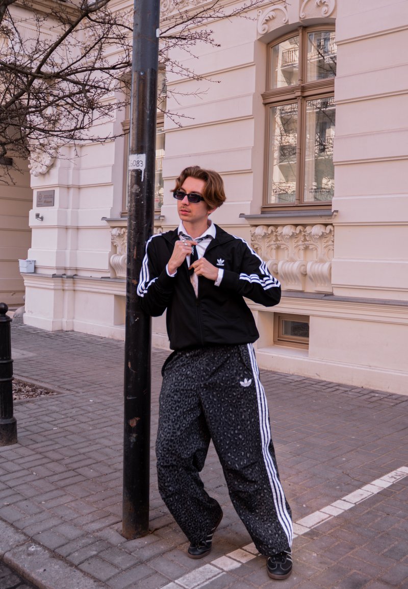 Junge Person in schwarzem Adidas-Trainingsanzug und Sonnenbrille passt ihre Krawatte an, während sie neben einer Straßenlaterne auf einem Bürgersteig mit verzierter Gebäudefassade in der Stadt steht.