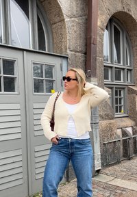 Kremasta, skraćena kardigana preko bijele majice, u kombinaciji s plavim trapericama. Sadrži sunčane naočale, torbu preko ramena i pozadinu od teksturirane kamenite zgrade.