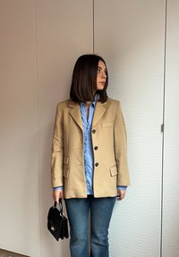 Žena s hnedými vlasmi po ramená, oblečená v béžovom saku, modrej košeli a džínsoch, drží čiernu kabelku, stojí pred textúrovanou stenou.