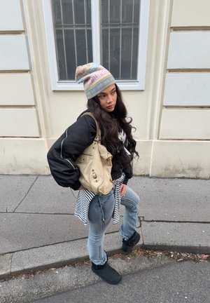 Joven con el pelo largo que lleva un gorro de punto con estampado en tonos pastel, chaqueta negra, camiseta de rayas anudada a la cintura, vaqueros, botas negras y lleva un bolso beige por la calle.