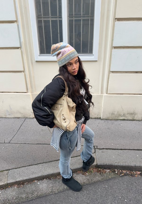 Jauna moteris su ilgais plaukais, dėvinti pastelinės spalvos raštuotą kepuraitę, juodą striukę, juostuotą marškinėlį, surištą ties liemeniu, džinsus, juodus batus, nešiojanti smėlio spalvos krepšį gatvėje.