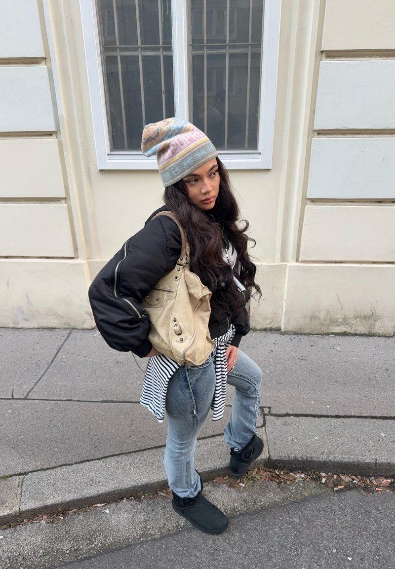 Νεαρή γυναίκα με μακριά μαλλιά, που φοράει παστέλ σκουφάκι με σχέδιο, μαύρο μπουφάν, ριγέ πουκάμισο δεμένο στη μέση, τζιν, μαύρες μπότες και κρατάει μπεζ τσάντα στο δρόμο.