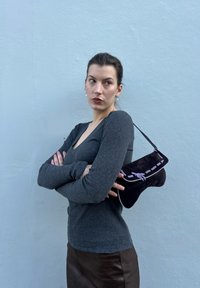 Sac à main en daim noir avec coutures violettes, design incurvé et sangle courte, tenu contre un haut gris à manches longues et une jupe marron.