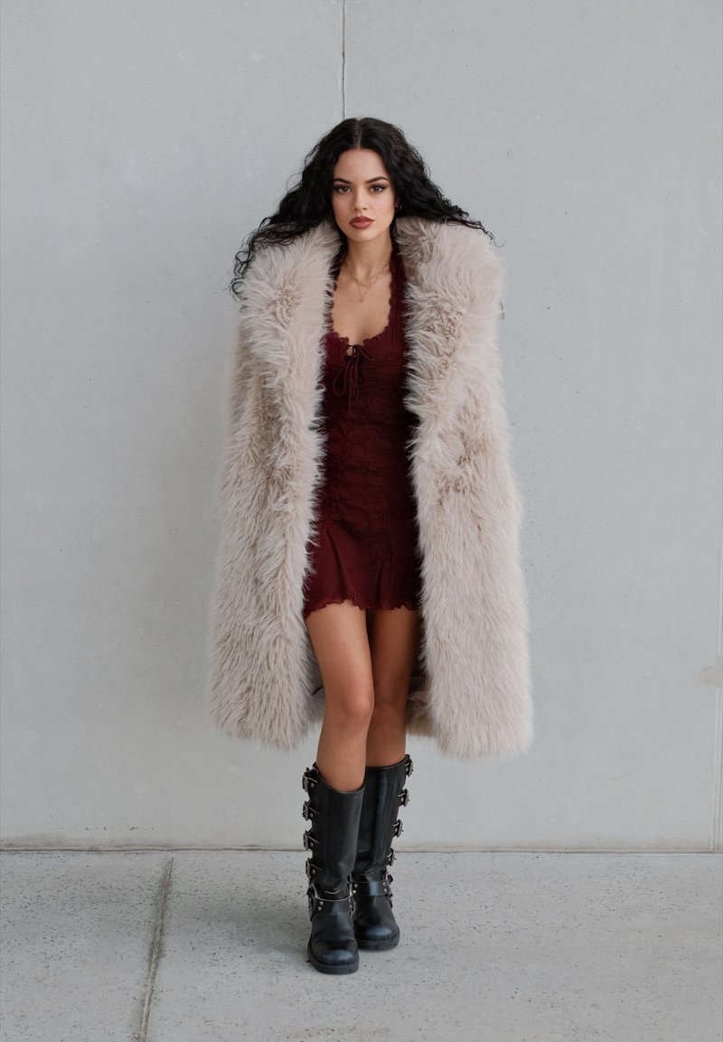 A long, fluffy beige coat worn over a burgundy dress featuring lace detailing. Black knee-high boots with metallic accents. A smooth wall backdrop.