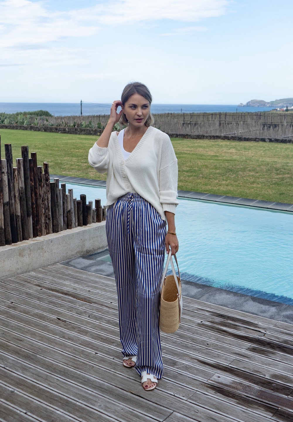Cardigan bianco lavorato a maglia, top bianco, pantaloni a gamba larga a righe blu e bianche, sandali bianchi, tiene in mano una borsa tessuta strutturata, in piedi accanto a una piscina.