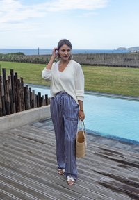 Cardigan bianco lavorato a maglia, top bianco, pantaloni a gamba larga a righe blu e bianche, sandali bianchi, tiene in mano una borsa tessuta strutturata, in piedi accanto a una piscina.