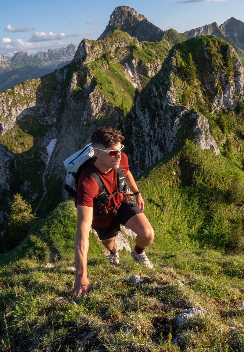 Young man wearing sunglasses and a backpack climbing a steep grassy mountain ridge with rugged peaks in the background.