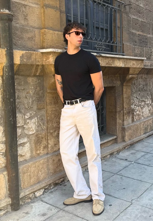 Homme portant un t-shirt noir, un pantalon blanc, des chaussures beiges et des lunettes de soleil, debout les mains derrière le dos contre un mur en pierre sur le trottoir.