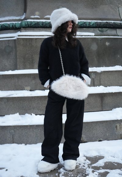 Mujer con un gorro peludo blanco, suéter negro, pantalones anchos negros y zapatos blancos, sosteniendo un bolso peludo blanco en unos escalones al aire libre cubiertos de nieve.