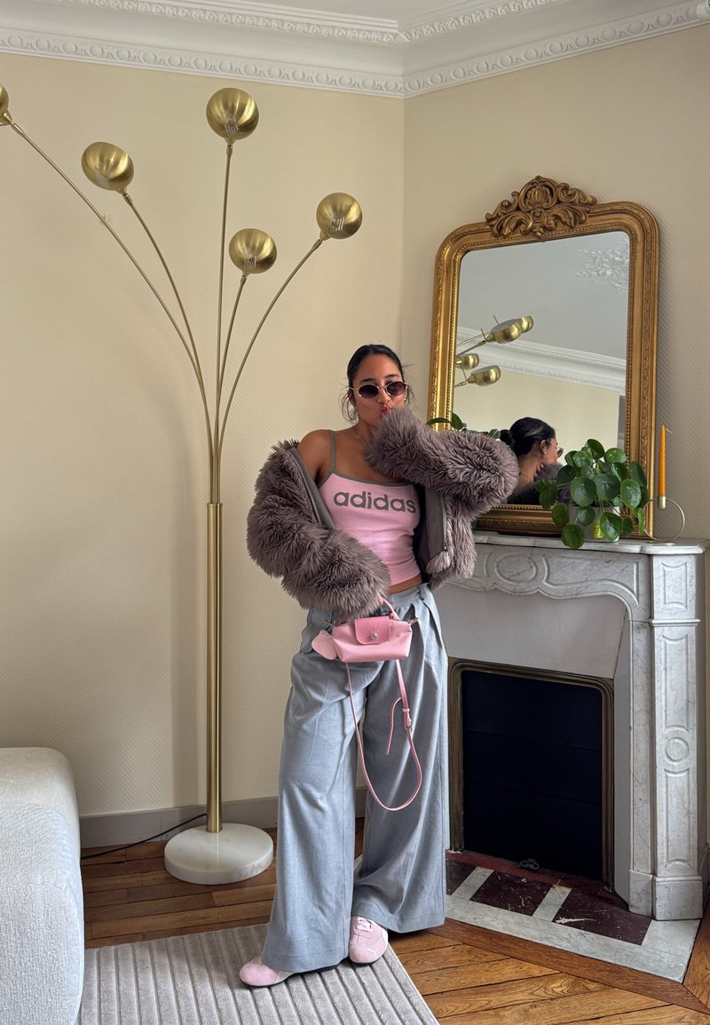 Femme en veste en fourrure oversize, haut Adidas rose, pantalon large gris et chaussures roses posant à côté d'une cheminée en marbre avec miroir encadré en or et plante.