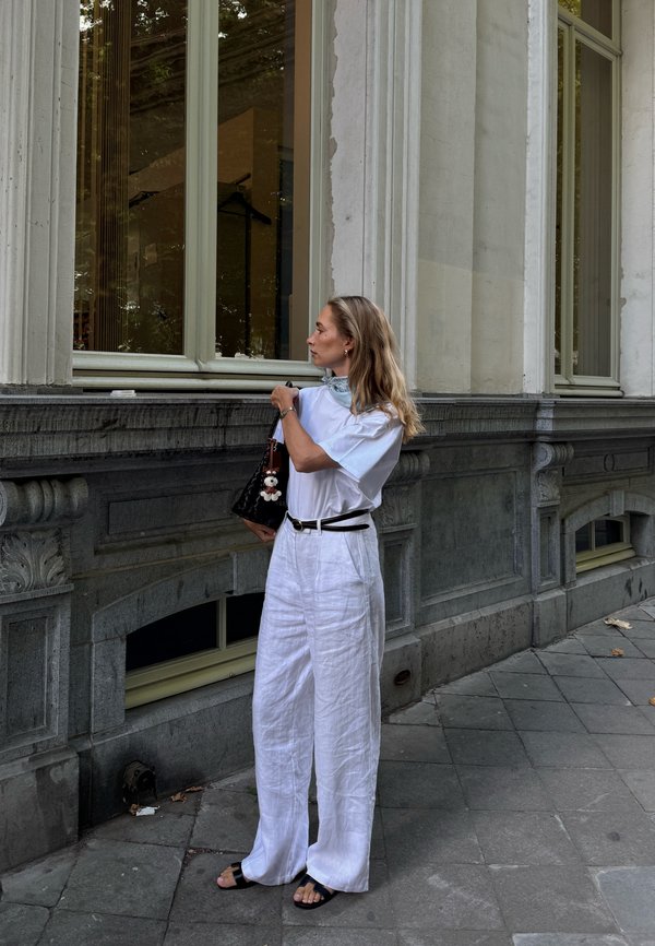 Pantaloni larghi bianchi in lino, t-shirt bianca loose, cintura nera e borsa nera con charm, in piedi contro un muro di pietra vicino a grandi finestre.