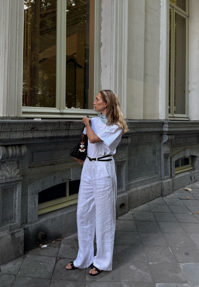 Bianco pantaloni larghi in lino, t-shirt bianca ampia, cintura nera e borsa nera con charm, in piedi contro un muro di pietra vicino a grandi finestre.