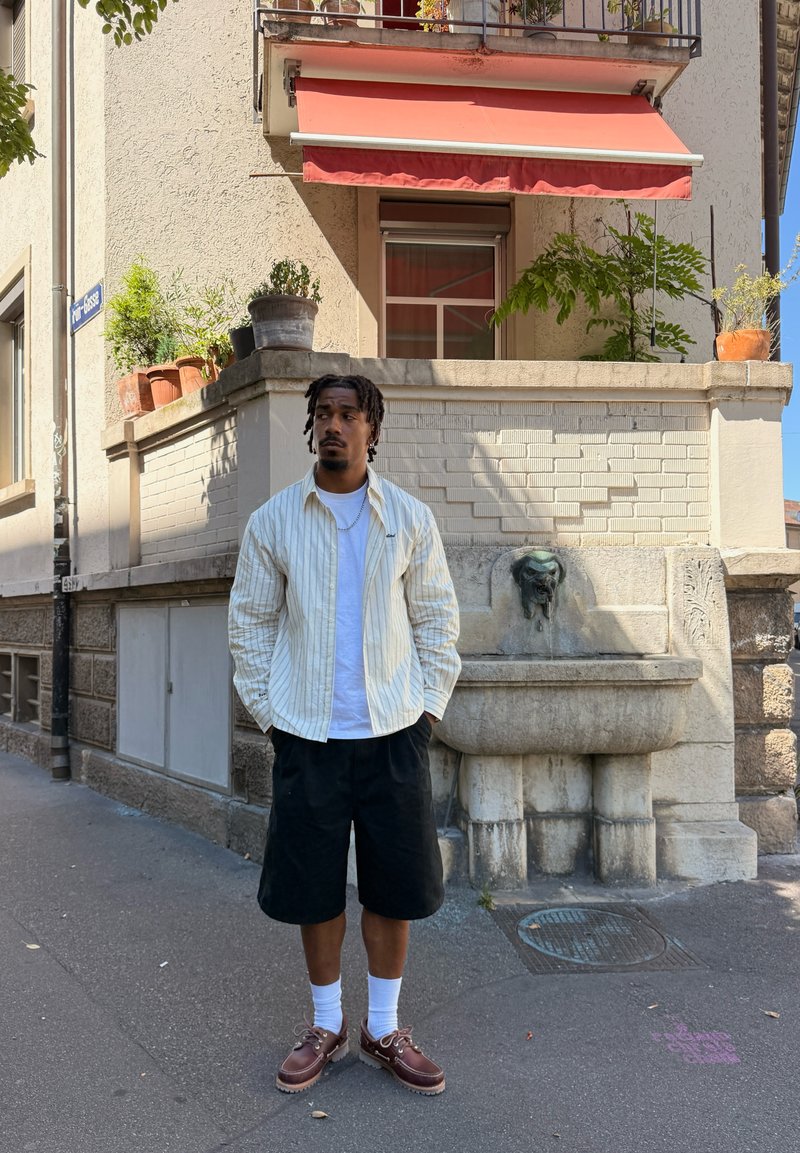 Uomo con le mani in tasca in piedi sulla strada vicino a un edificio beige con una tenda rossa, piante in vaso e una fontana di pietra con un beccuccio a forma di testa di leone.