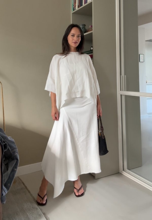 Witte linnen top met oversized mouwen, gecombineerd met een witte asymmetrische rok en zwarte teenslippers. Houdt een zwarte handtas vast.