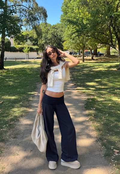 Top corto blanco, suéter de punto crema drapeado, pantalones de pierna ancha azul marino, zapatos de plataforma beige, sosteniendo un bolso de color claro en un parque.