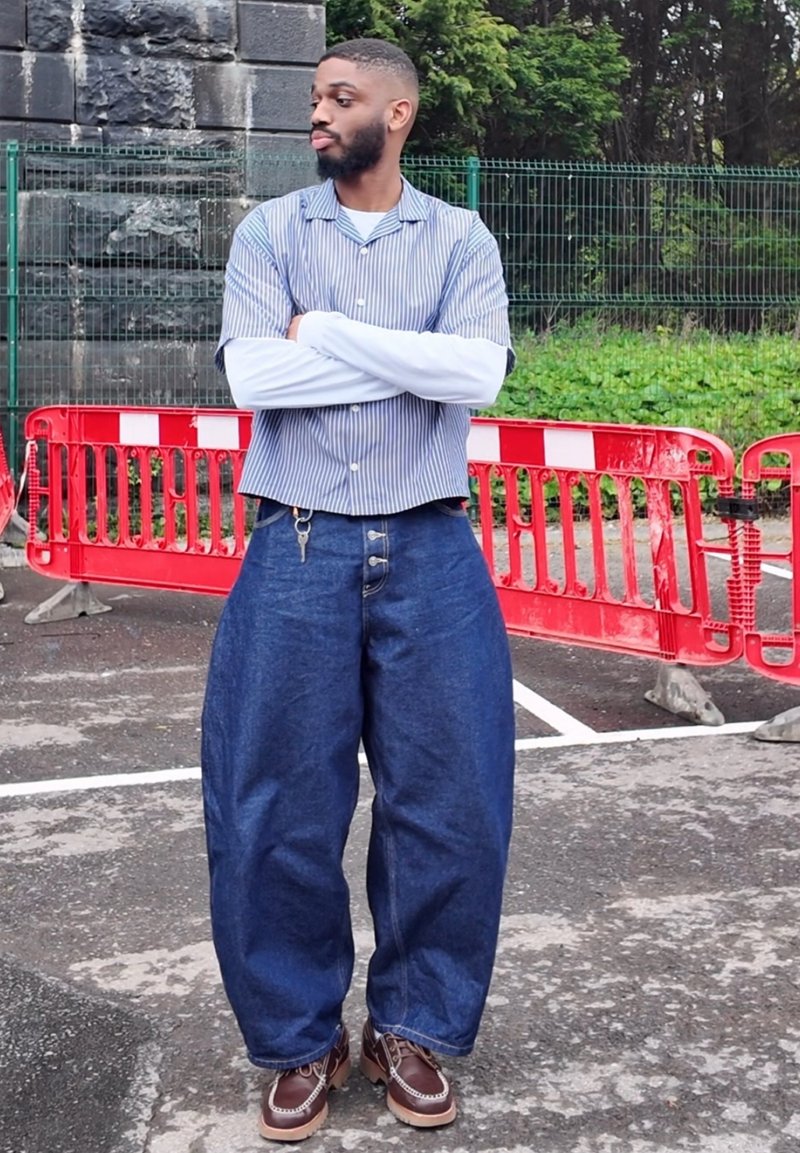 Homme debout, bras croisés, portant des chemises superposées, un jean large et surdimensionné et des chaussures marron près de barrières rouges et un mur en pierre.