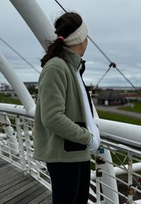 Veste en polaire de couleur vert clair, avec une poche noire, portée par-dessus un sweat-shirt gris. Des leggings noirs et un bandeau complètent la tenue.