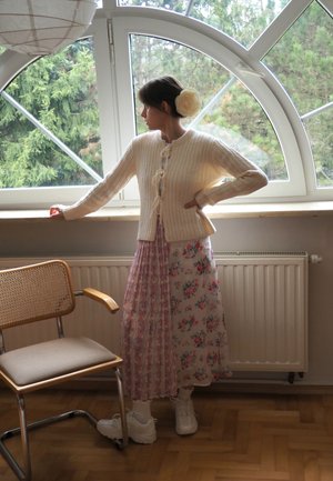 Cárdigan blanco acanalado con lazo en el frente, combinado con una falda midi floral en rosa y blanco. Zapatillas blancas, silla de madera y grandes ventanales.