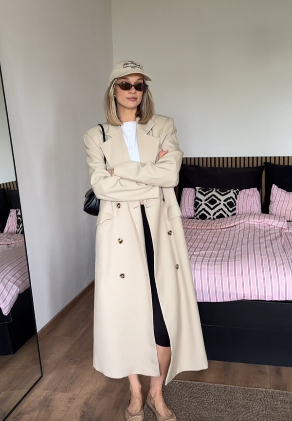 Mujer con abrigo largo beige, camisa blanca, falda negra, gorra beige, gafas de sol, de pie con los brazos cruzados en un dormitorio con ropa de cama a rayas rosas.