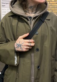 Palton oversized verde măsliniu, cu închideri cu nasturi, purtat peste un hanorac bej, cu o curea neagră și tatuaje vizibile pe mână.