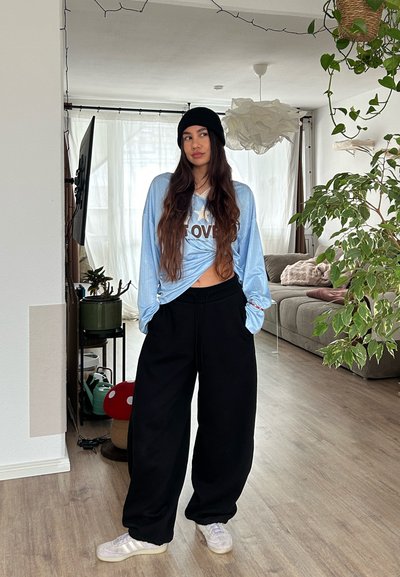 Joven con gorro negro, camisa azul claro de manga larga anudada con el texto "LOVE", pantalones anchos negros y zapatillas blancas, en interiores cerca de plantas y un sofá.