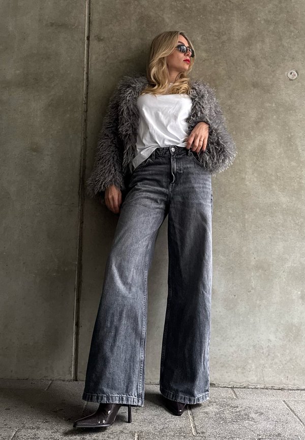 Grijze jas van imitatiebont, wit t-shirt, grijze wijde jeans met hoge taille en puntige donkere hakken. Model draagt een zonnebril en leunt tegen een betonnen muur.