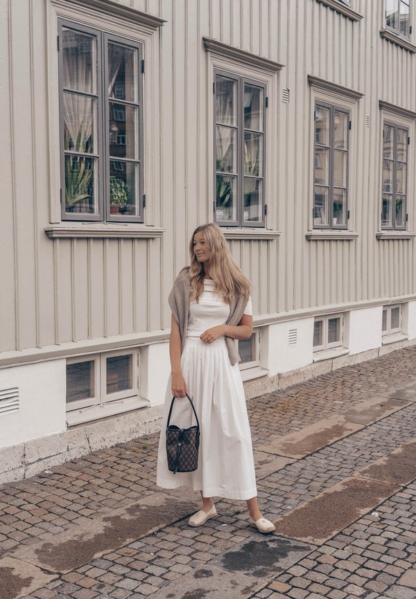Femme en robe blanche et châle beige debout dans une rue pavée à côté d'un bâtiment en bois beige avec plusieurs fenêtres et des plantes en pot à l'intérieur.