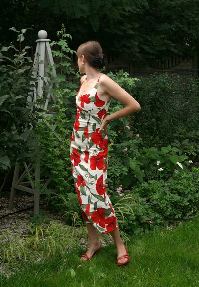 Vestido midi floral con patrones de amapolas rojas sobre un fondo blanco. Presenta tirantes finos, silueta ajustada y dobladillo a la altura de la rodilla. Sandalias rojas.